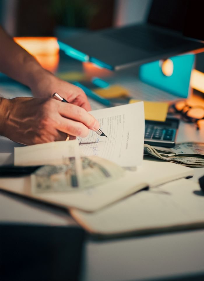 Services A hand signing financial documents on a desk with cash and a calculator, signifying finance management.